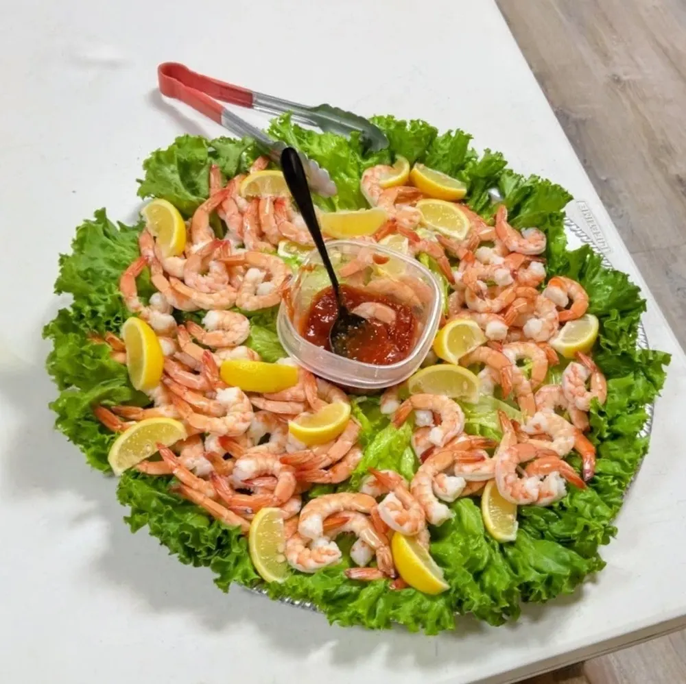 A catering platter of shrimp cocktail on a bed of lettuce, with lemon wedges and a cup of cocktail sauce in the middle.