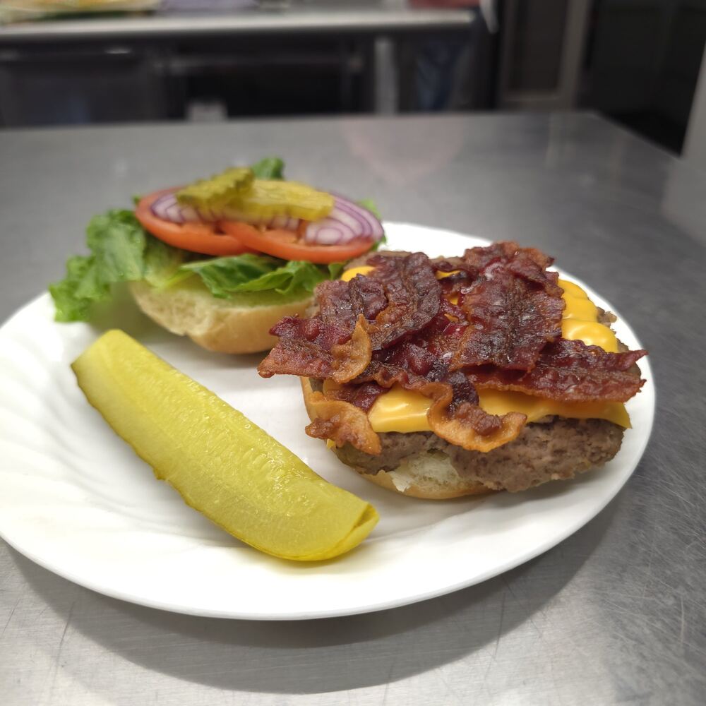 Bacon cheeseburger served open-faced on a white plate: a beef patty with melted American cheese and crisp bacon, the top bun with lettuce, tomato, red onion and pickles, and a pickle spear.