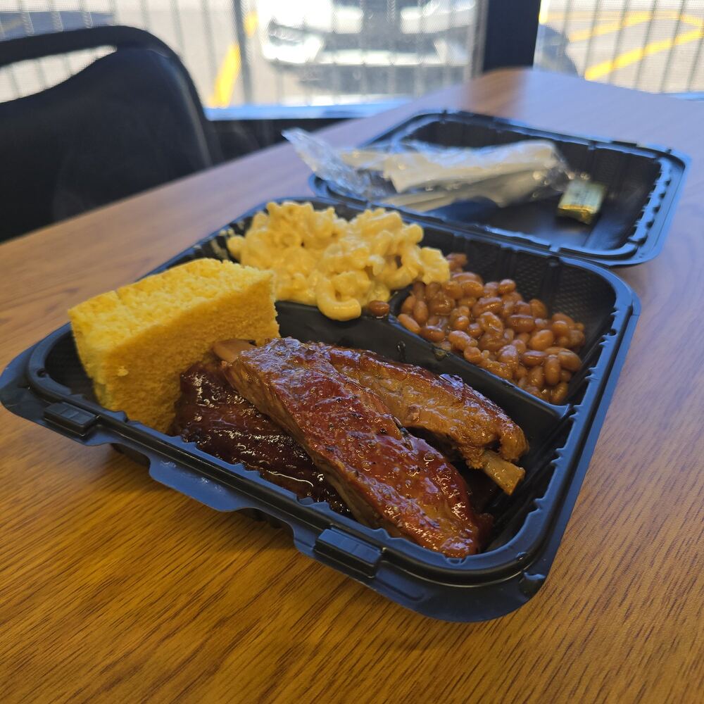BBQ ribs glazed in sauce in a to-go box, with a square of cornbread, mac and cheese, and baked beans.