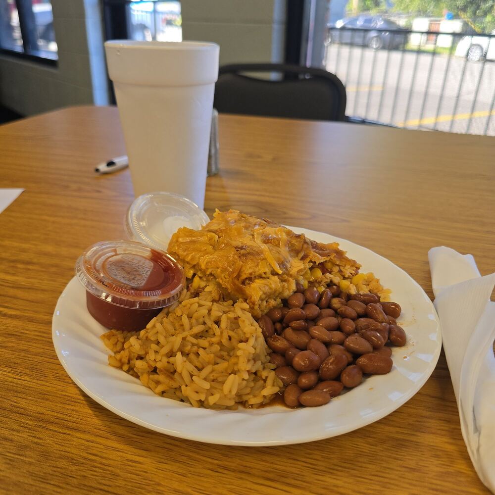 Chicken enchilada casserole topped with melted cheese, with Spanish rice, pinto beans and cups of salsa and sour cream on a white plate.