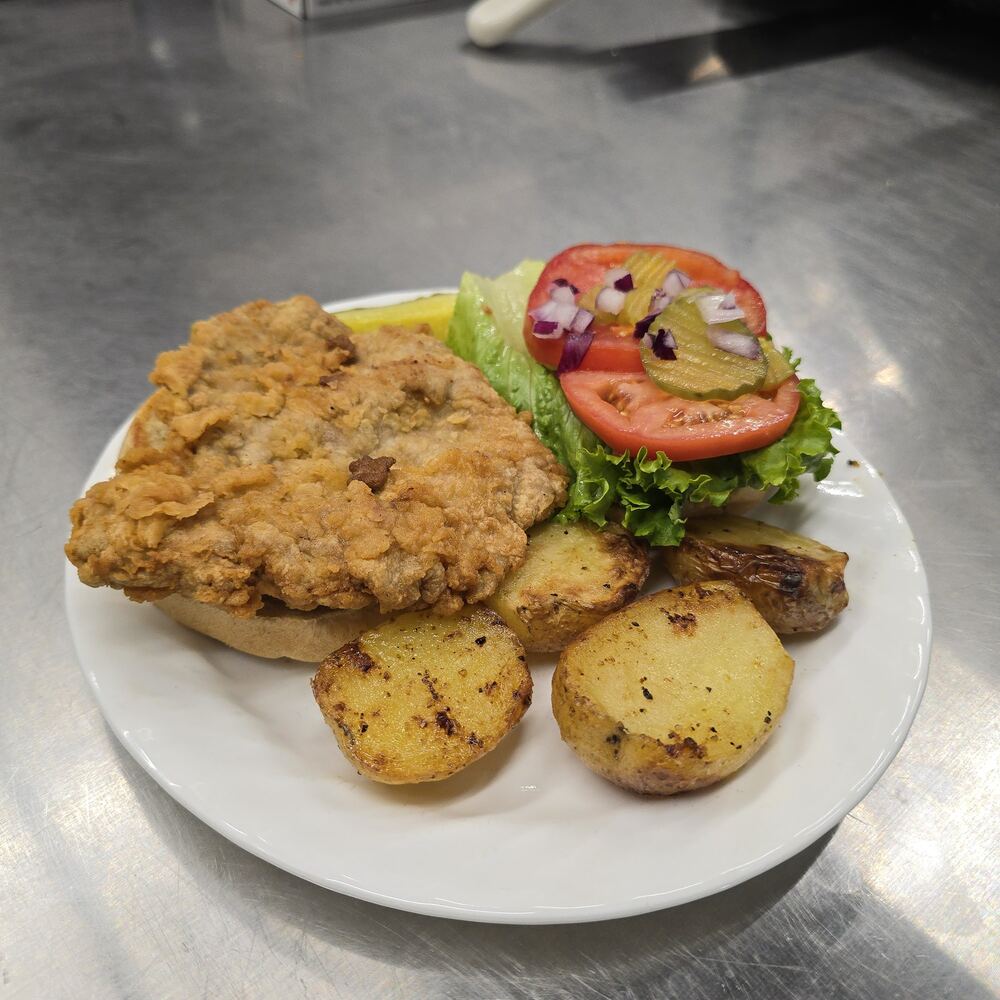 A breaded pork tenderloin on an open bun, with lettuce, tomato slices, pickles and diced red onion, and oven baked potato wedges on a white plate.