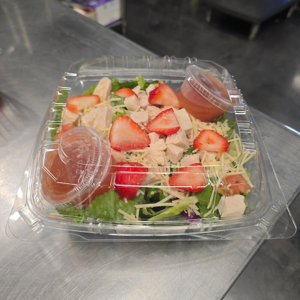 Strawberry chicken salad in a clear to-go container: greens topped with diced chicken, sliced strawberries, tomato and shredded parmesan, with two cups of dressing on the side.